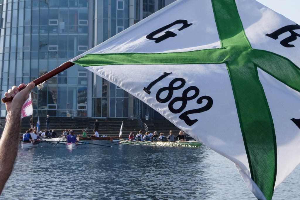Die Vereinsflagge der Rudergesellschaft Germania Kiel wird von einem Arm gehalten, im Hintergrund sind mehrere Ruderboote zu sehen.