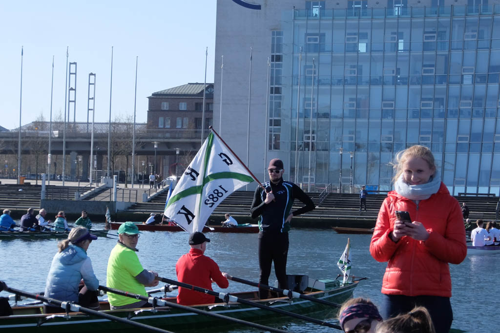 Mann steht in einem Ruderboot und hält eine Vereinsflagge in der Hand, neben ihm steht eine Frau in einem anderen Ruderboot und tippt auf ihrem Handy
