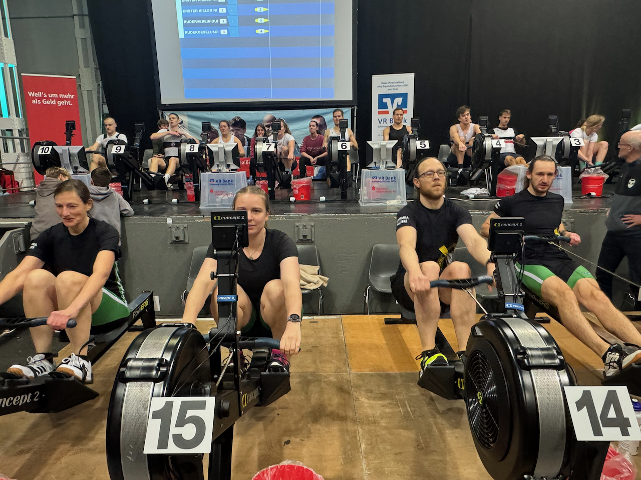 Zwei Frauen und zwei Männer sitzen nebeneinander auf Ruderergometern vor einer Bühne. Auf der Bühne rudern weitere Personen auf einem Ergometer.
