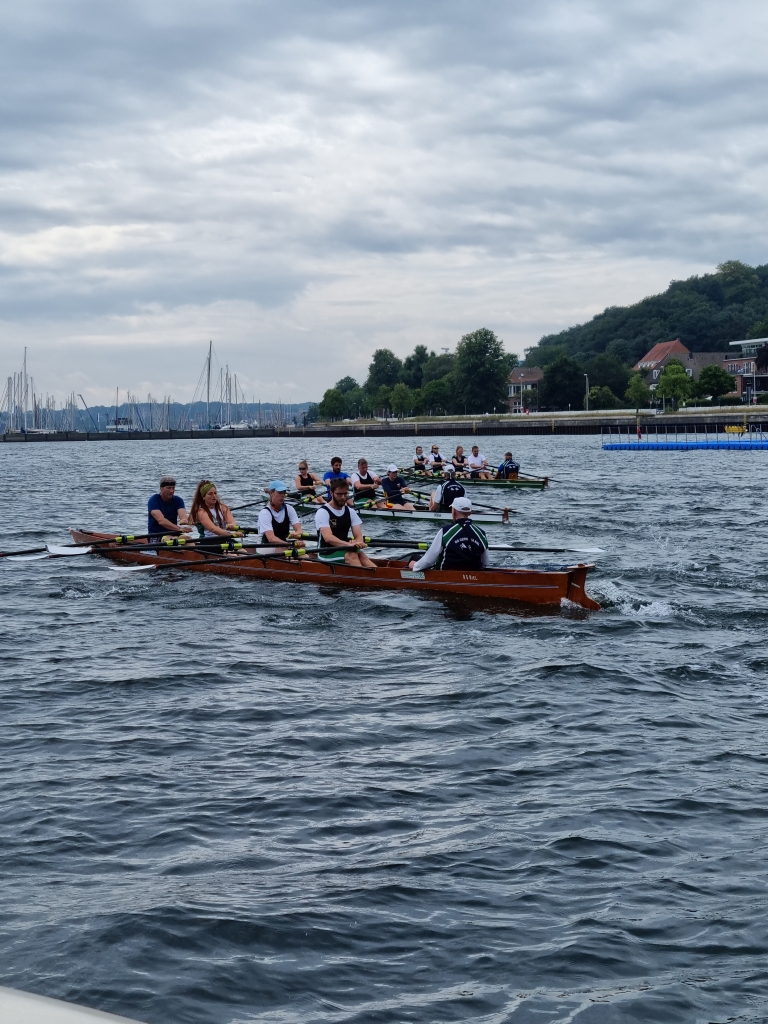 Mehrere Personen in drei Ruderbooten in einem Rennen vor einem Hafen mit Segelbooten 