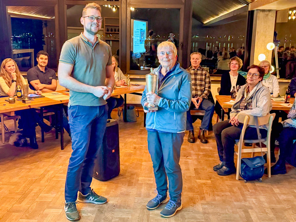 Eine ältere Frau hält einen Pokal in der Hand den sie von dem neben ihr stehenden jungen Mann erhalten hat. Im Hintergrund sitzen Personen und schauen interessiert zu.