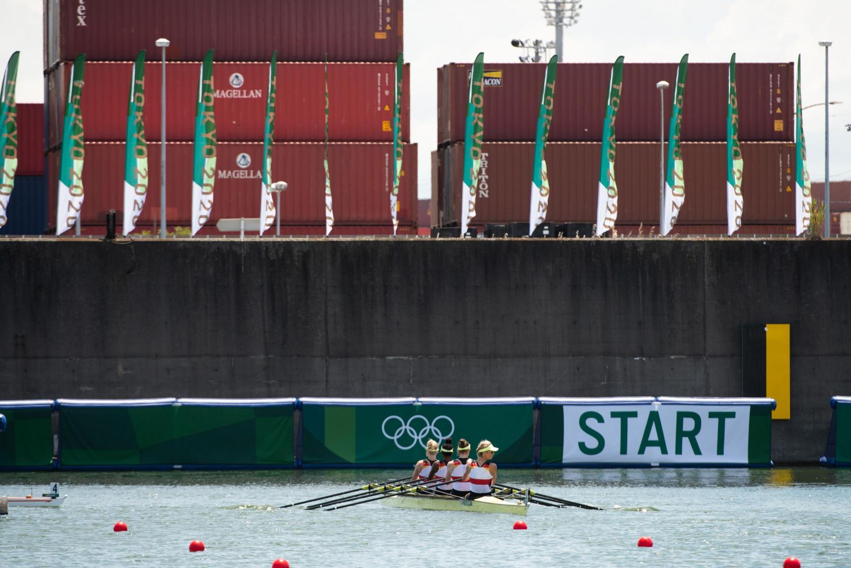 Vier Frauen in einem gelben Ruderboot vor einer Mauer beschrieben mit Start und den Olympischen Ringen obenauf Fahnen und Container