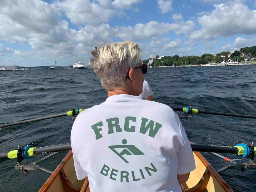 Ein Ruderer mit der Aufschrift "FRCW Berlin" auf dem Rücken blickt vom Ruderboot über die Kieler Förde zum Westufer.