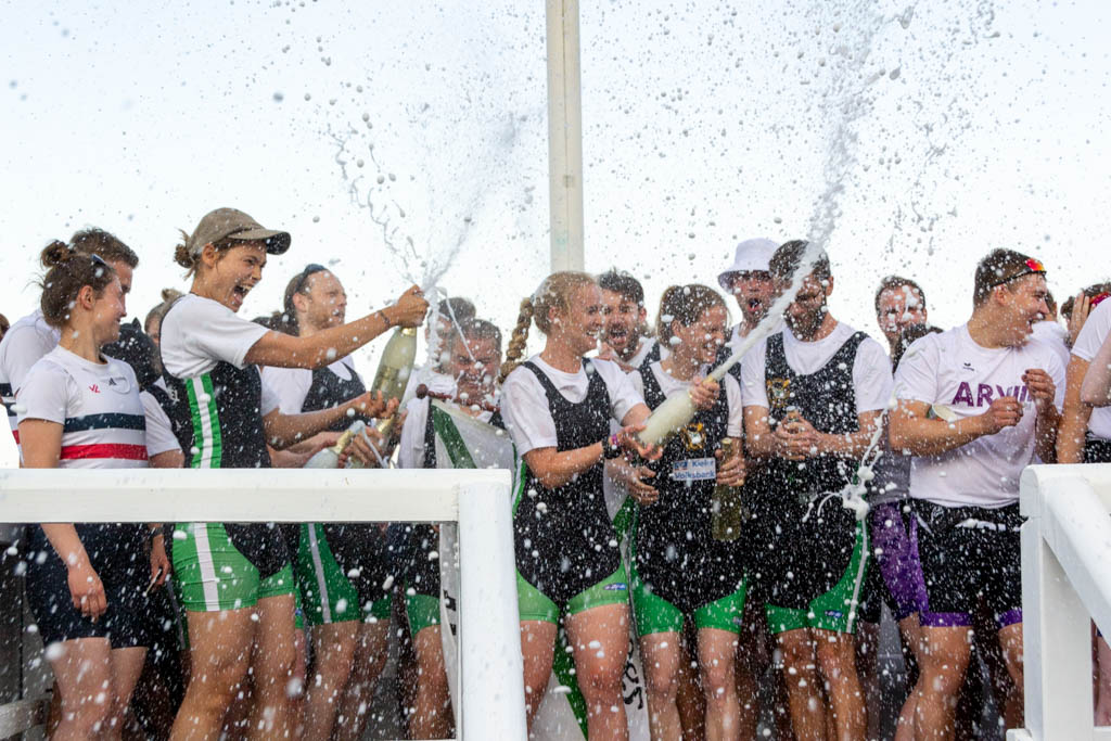 Mehrere Personen in weiß-grün.schwarzer Sportkleidung freuen sich bei der Siegerehrung des Stadtachterrennens über den gewonnen Pokal und verspritzen Sekt aus Flaschen.