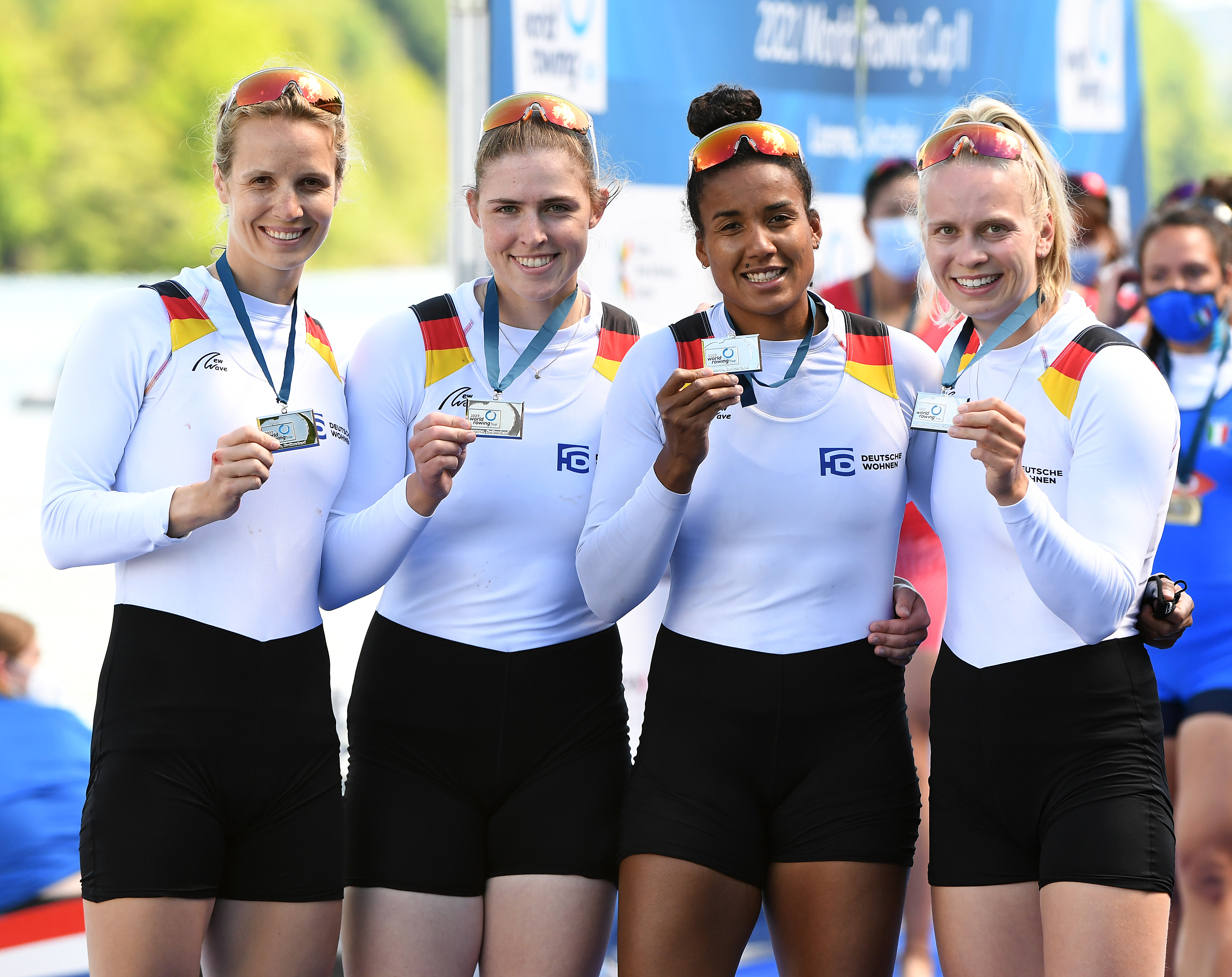 Vier Frauen im Sporttrikot stehen nebeneinander vor einer Plakatwand und halten Silbermedaillen in der Hand