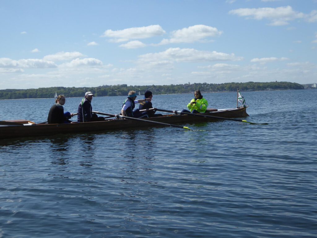 ein Ruderboot schwimmt auf dem Wasser mit fünf Personen darin, im Hintergrund eine Baumreihe am Ufer