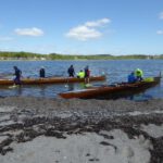Zwei Ruderboote liegen am Ufersaum eines Gewässers unds mehrere Personen sind in den Booten oder drumherum