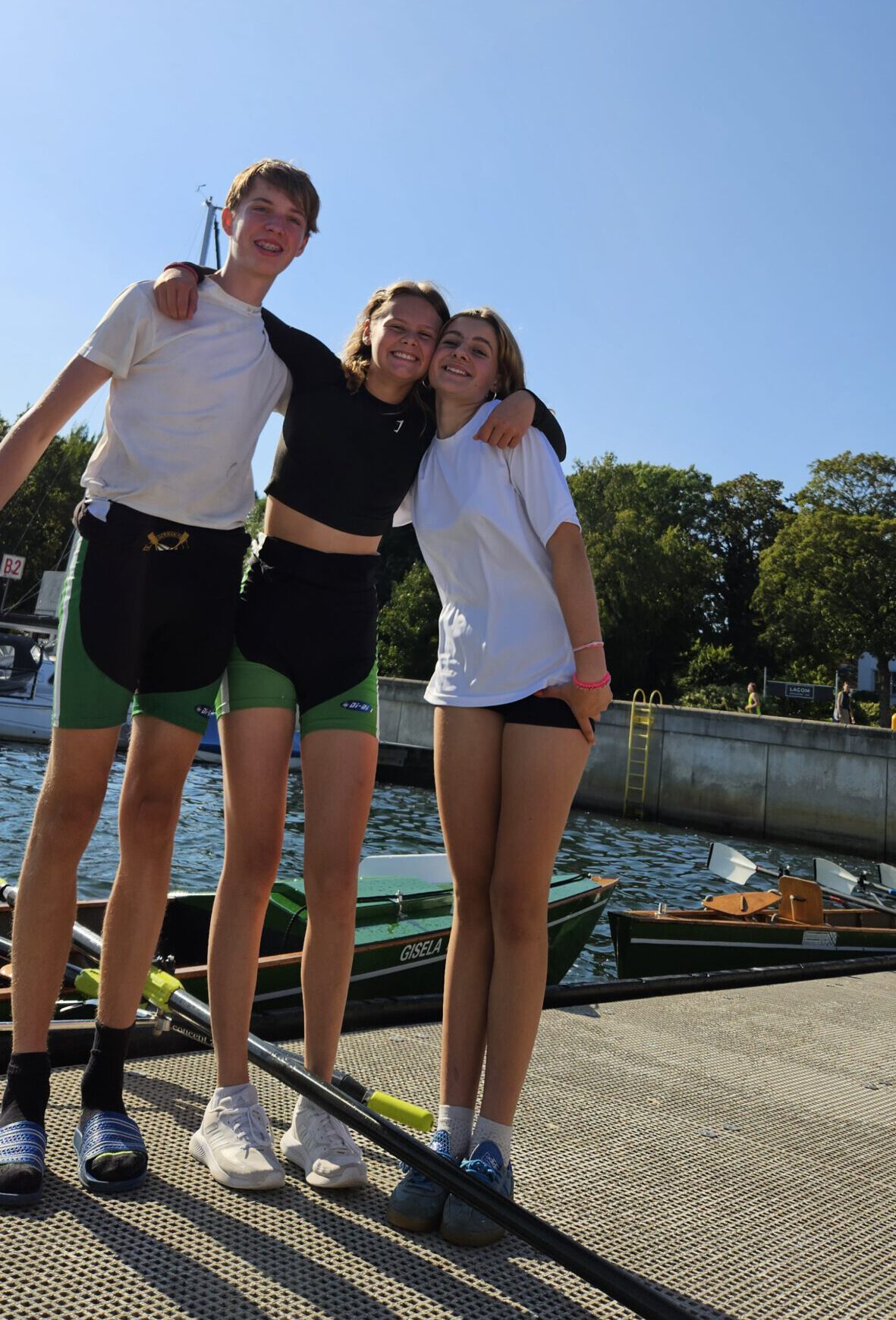 Ein Junge und zwei Mädchen stehen auf einem Bootssteg vor zwei Ruderbooten und umarmen sich.