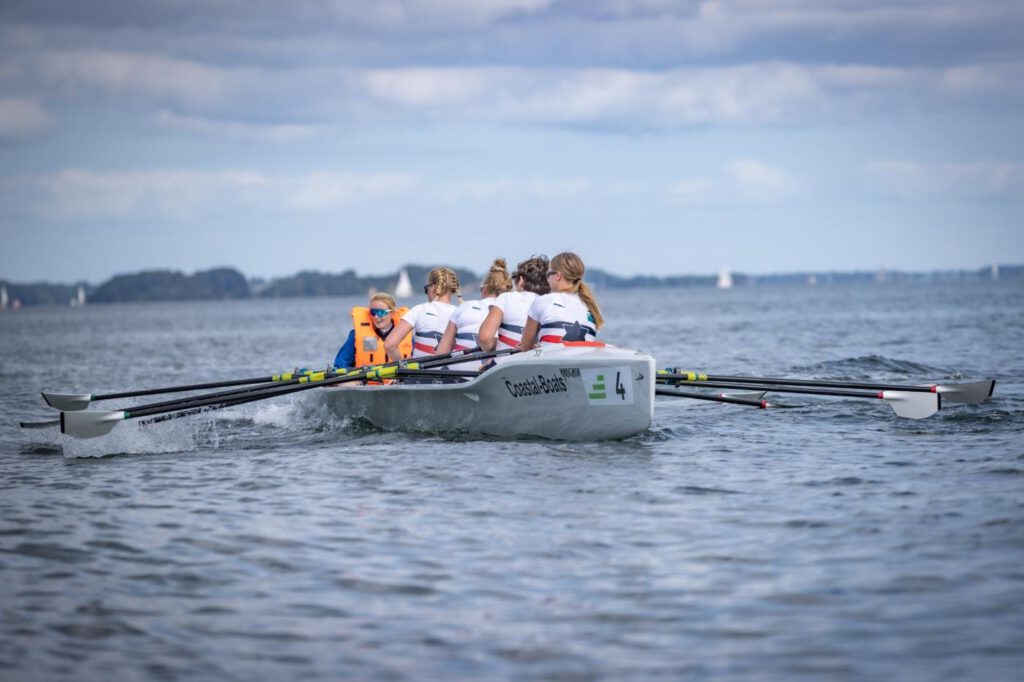 Vier Frauen sitzen hintereinander in einem Ruderboot und vor ihnen die Steuerfrau mit einer orangen Rettungsweste