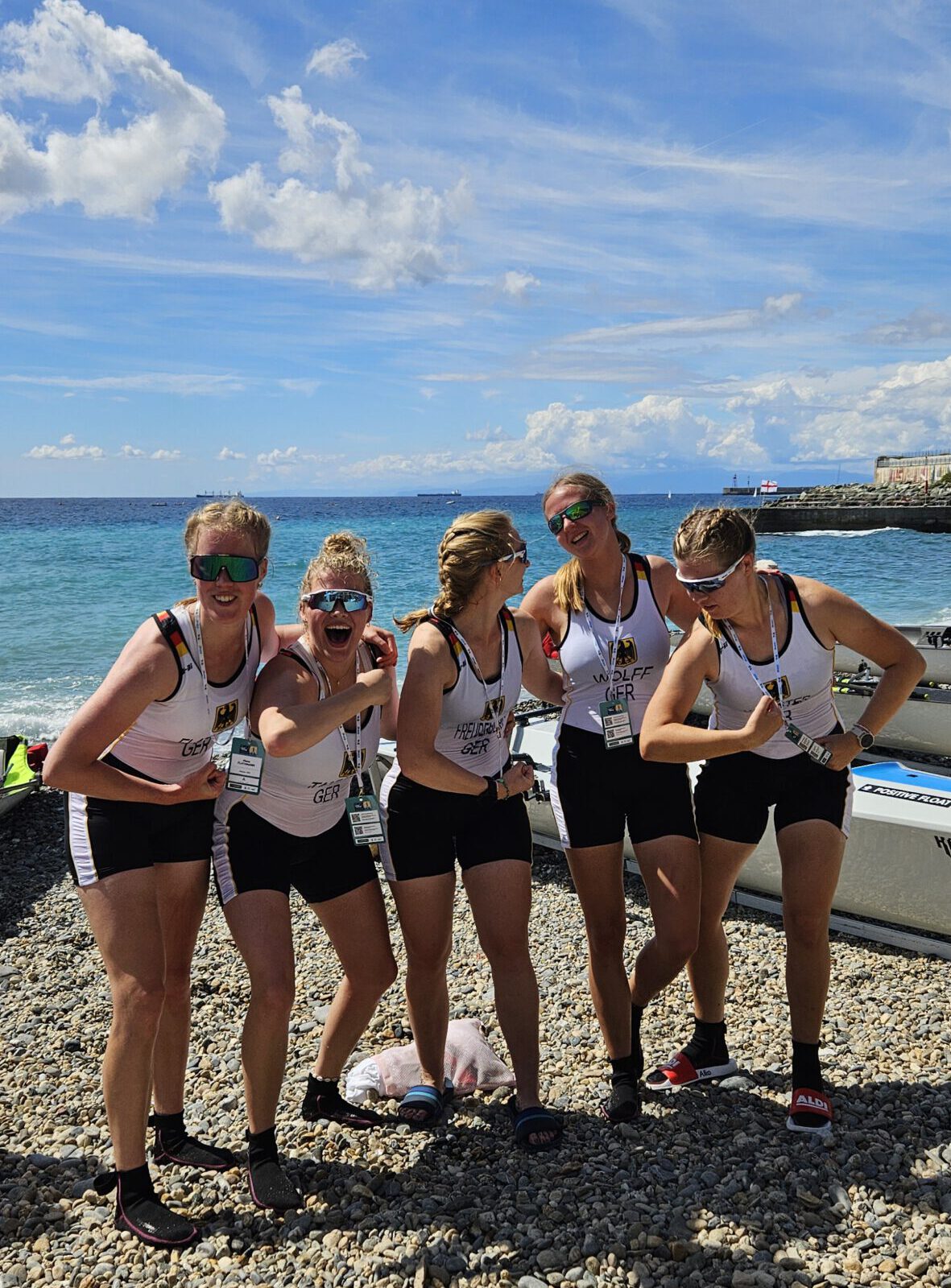 Fünf Frauen in Sportkleidung stehen lachend und jubelnd nebeneinander am Strand