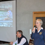 Eine Frau steht und spricht in ein Mikrofon, neben ihr sitzt ein Mann vor einer Leinwand auf die ein Bild projiziert wird.