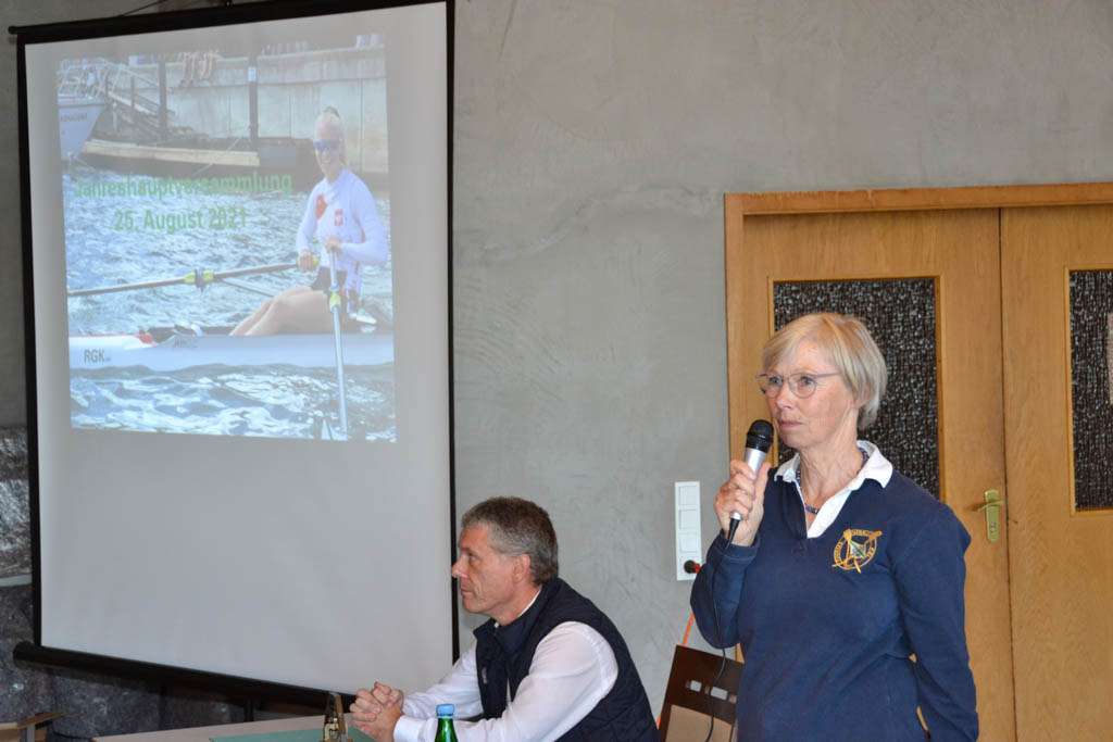 Eine Frau steht und spricht in ein Mikrofon, neben ihr sitzt ein Mann vor einer Leinwand auf die ein Bild projiziert wird.